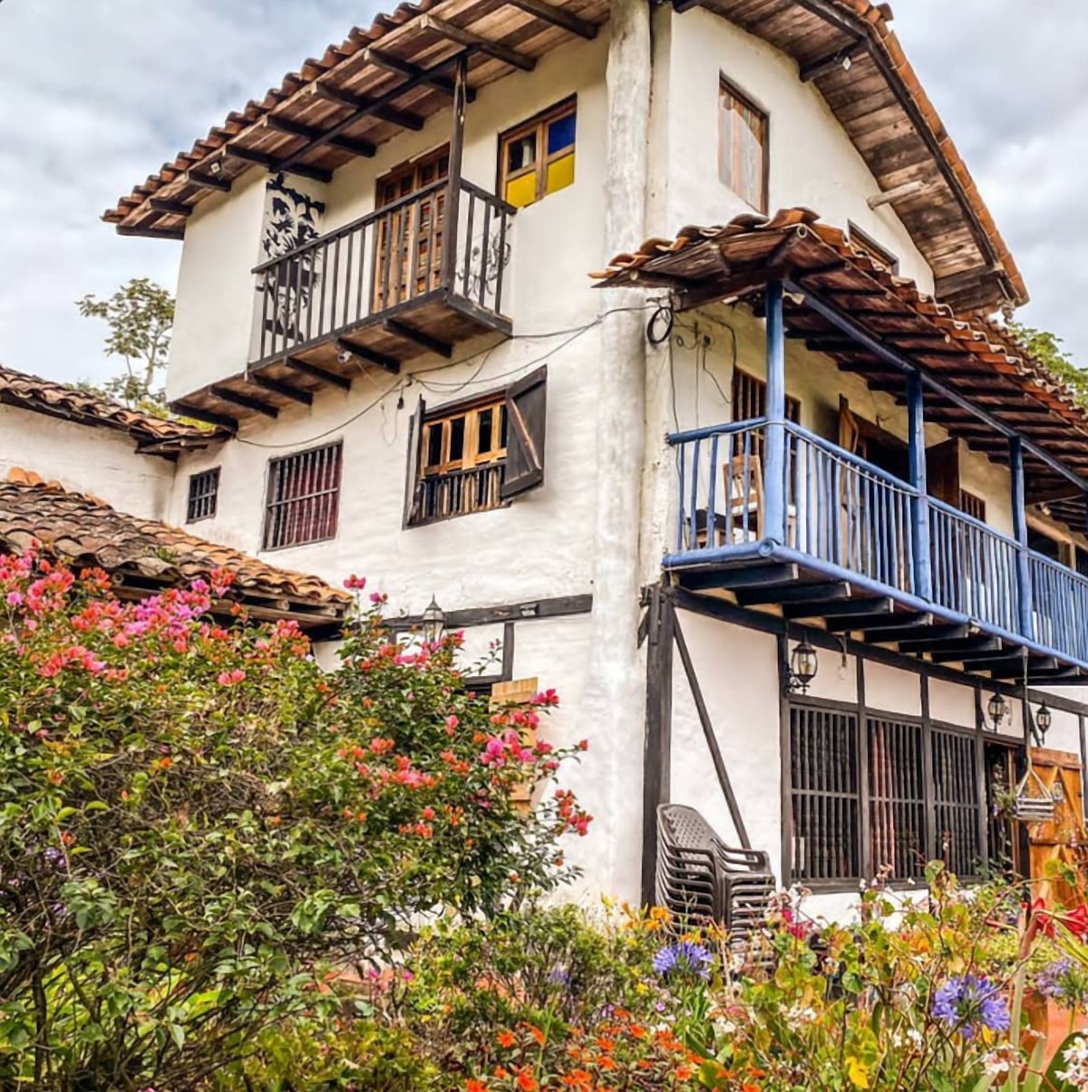 Casa de dos pisos de paredes blancas y madera, con balcones de madera azul y un jardín florecido en primer plano