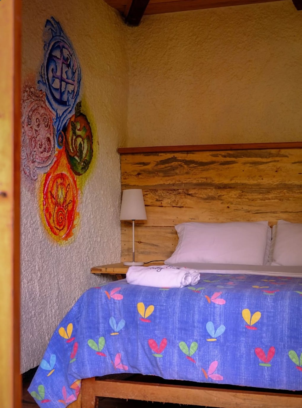 Cama doble con cobija azul estampada de flores, cabecero de madera y un mural de círculos de colores en la pared