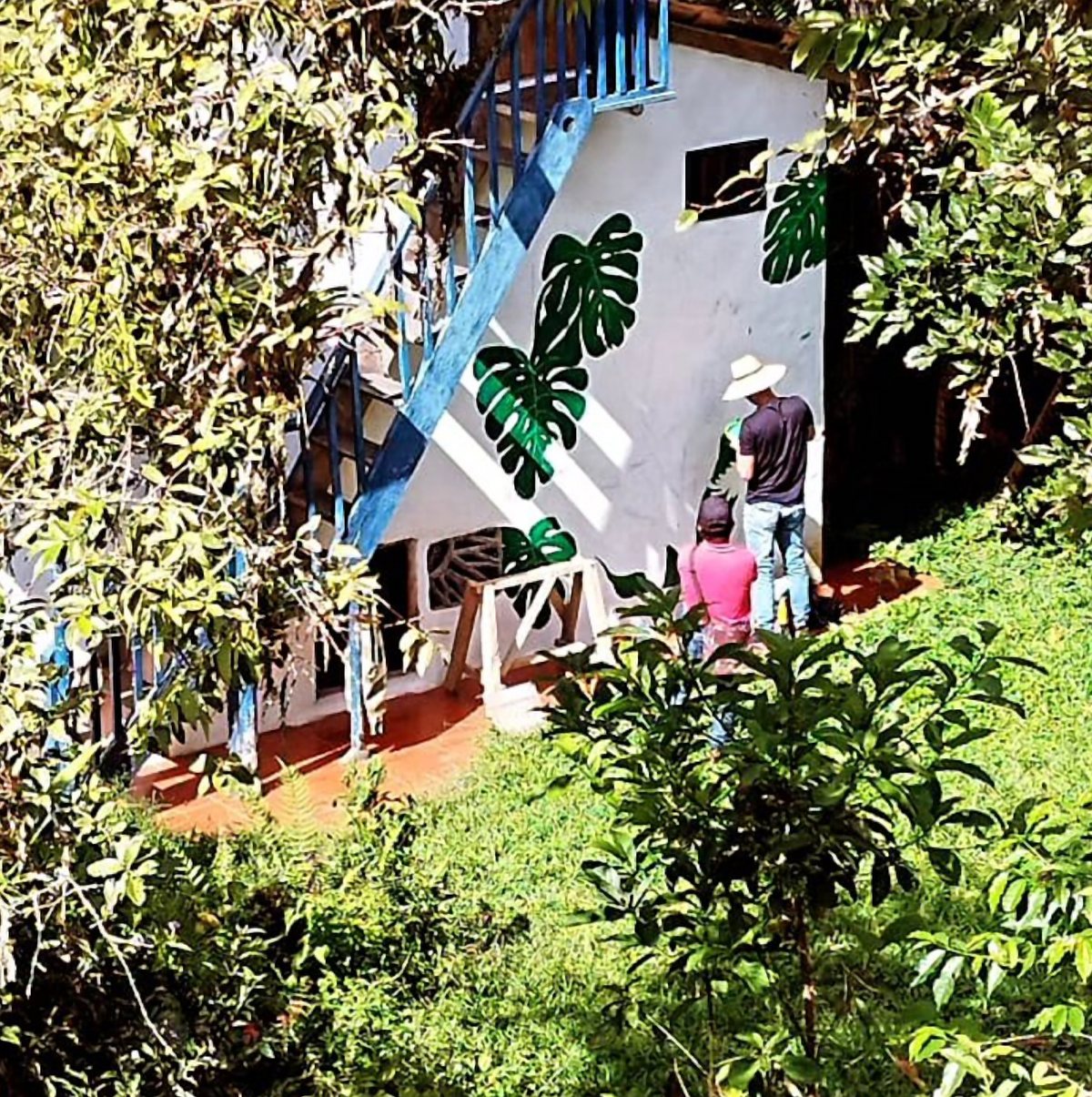 Pared blanca con un mural de hojas de monstera junto a una escalera azul, entre vegetación