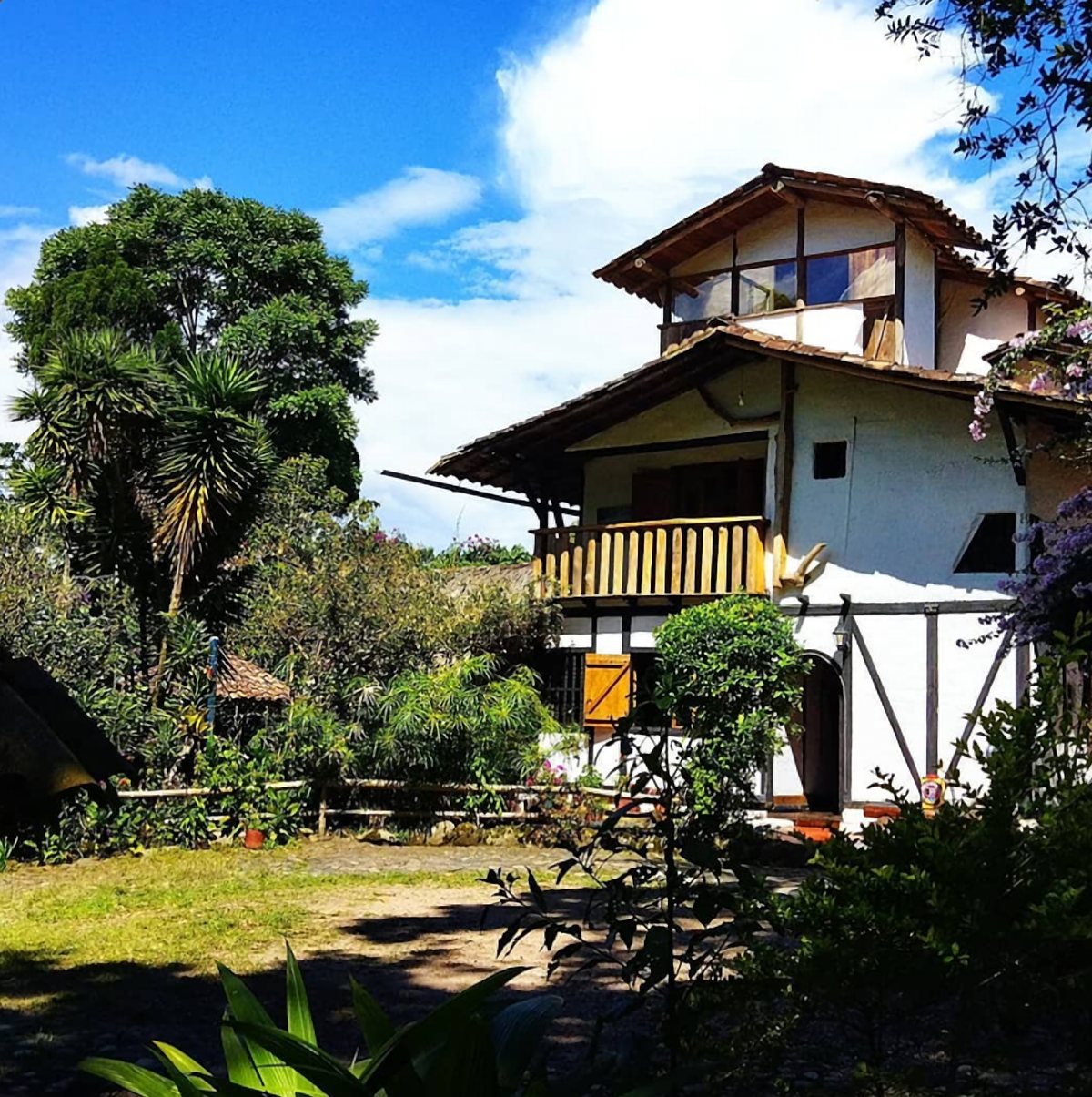Casa de dos pisos con balcón de madera y tejas de barro bajo un cielo azul, frente a un patio con jardín