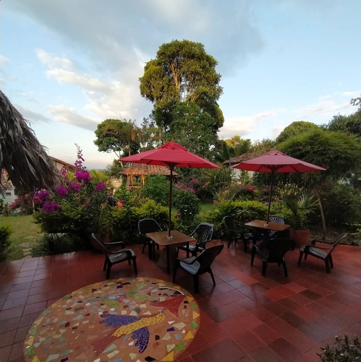 Terraza de baldosas con sombrillas rojas, mesas y un mosaico de colibrí en el piso, frente al jardín